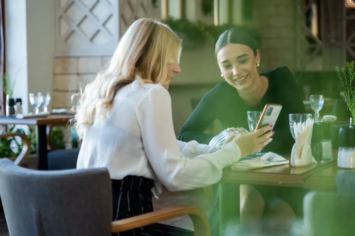 QR menu εμπειρία πελάτη: δύο πελάτισσες σε τραπέζι εστιατορίου βλέπουν χαμογελαστές το ψηφιακό μενού στο κινητό